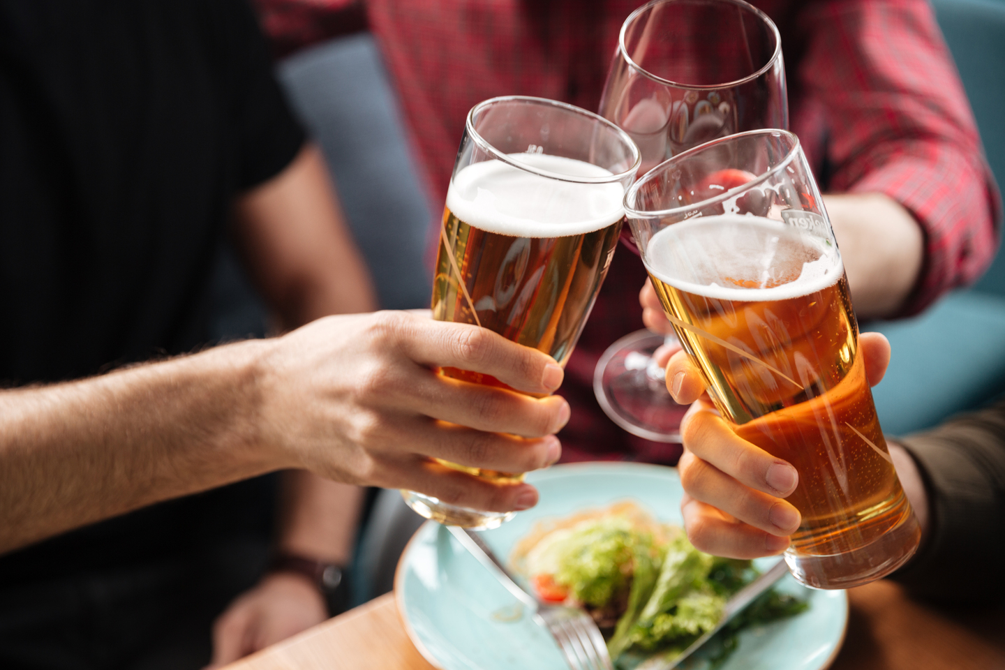 Cropped photo of young friends sitting in cafe while drinking alcohol. Focus on glasses of beer.